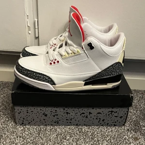 Nike air jordan 3 cement - Säljer ett par klassiska Nike Air Jordans i vitt med grå och svarta detaljer. Skorna har en unik mönstrad design runt tån och hälen, samt röda accenter. Perfekta för sneaker-entusiaster som vill ha en stilren look.