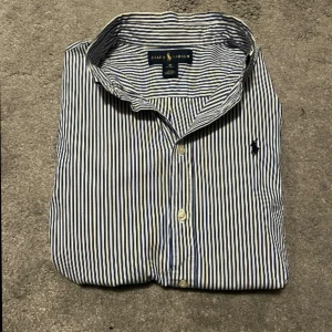 Randig skjorta från Ralph Lauren - Snygg blå- och vitrandig skjorta från Ralph Lauren med klassisk krage och broderad logga på bröstet. Skjortan har långa ärmar och knappar Stl xs