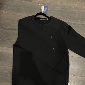 Svart tröja från Ralph Lauren - Säljer en stilren svart tröja från Ralph Lauren med det klassiska broderade logotypen i blått på bröstet. Tröjan har långa ärmar och en rund halsringning. Perfekt för en avslappnad och snygg look.