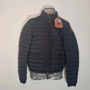 Svart dunjacka från Parajumpers - Säljer en svart dunjacka från Parajumpers i superlätt material. Jackan har en dragkedja framtill och en hög krage. Den är dekorerad med ett märke på ärmen och har en quiltad design. Perfekt för kyliga dagar!