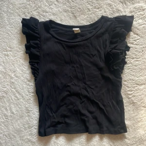 Svart ribbad topp med volangärmar - Snygg svart ribbad topp med volangdetaljer på ärmarna. Perfekt för att ge en extra touch till din outfit. Toppen är ärmlös och har en bekväm passform.