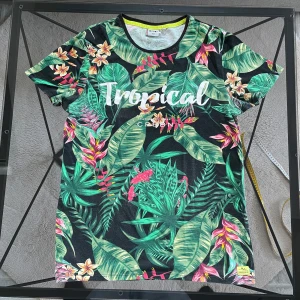 Tropisk t-shirt från Hailys - Snygg t-shirt från Hailys med tropiskt blommönster i grönt, rosa och gult. Texten 'Tropical' pryder framsidan. Perfekt för en somrig look!