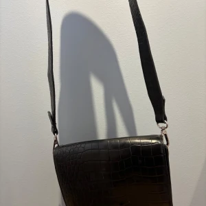 Svart väska - Snygg svart axelväska med krokodilmönster och justerbar rem. Väskan har en praktisk dragkedja och en ficka på framsidan för extra förvaring. Perfekt för att ge din outfit en stilfull touch.