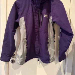Lila och grå jacka från Millet - Säljer  mörk lila/ grå jacka från Millet i Gore-Tex-material. Jackan har långa ärmar och flera praktiska dragkedjor. Perfekt för utomhusaktiviteter och skidåkning. Vind och  vattentät,  andningsbar design.