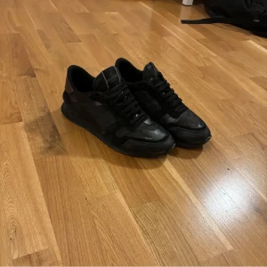 Valentino Garavani rockrunners svart - Snygga svarta sneakers från Valentino Garavani med ett diskret camouflage-mönster. Skorna har snörning och är tillverkade i en kombination av skinn och syntetmaterial. Perfekta för en stilren look.