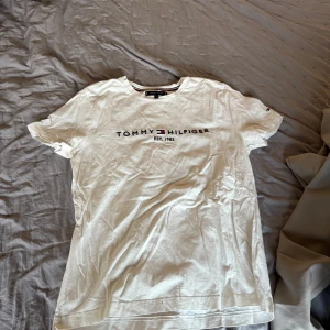Vit t-shirt från Tommy Hilfiger - Säljer en klassisk vit t-shirt från Tommy Hilfiger med tryck på bröstet. T-shirten har korta ärmar och rund halsringning. Perfekt för en avslappnad stil.