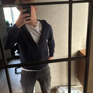 Mörkblå hoodie från Ralph Lauren - Säljer en snygg mörkblå hoodie från Ralph Lauren med dragkedja och det klassiska röda logotypen på bröstet. Perfekt för en avslappnad stil. Hoodien har långa ärmar och en bekväm passform.