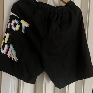 Svarta shorts med grafiskt tryck från Firefly - Coola svarta shorts från Firefly med färgglatt grafiskt tryck och dödskallemotiv. De har en elastisk midja med gula snören för justering. Perfekta för en avslappnad stil.