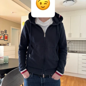 Mörkblå huvtröja från Moncler - Säljer en snygg mörkblå huvtröja från Moncler med dragkedja och ribbade muddar. Tröjan har en broderad logga på ärmen och vita samt röda detaljer vid ärmslut och krage. Perfekt för en stilren look!