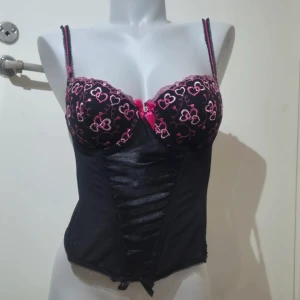 Svart korsett med hjärtmönster - Snygg svart korsett med rosa hjärtmönster och spetsdetaljer. Den har en vacker snörning framtill och en söt rosett mellan kuporna. Perfekt för en speciell kväll!
