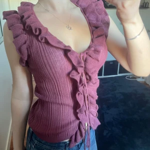 Vinröd ribbad topp med volangdetaljer - Säljer den trendiga ribbade toppen från Zara med volangdetaljer längs halsringningen och knytning framtill. Toppen är ärmlös och har en figurnära passform. Finns minimala slitningar på ett ställe, som var där redan vid köpet ❣️Inget  större man lägger märke till.