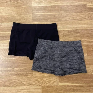Två par shorts i svart och grått - Säljer två par shorts, ett par i svart och ett par i gråmelerat. Båda är i en tight passform och tillverkade i ett stretchigt syntetmaterial. Perfekta för träning eller avslappnade dagar. De har en elastisk midja för extra komfort.