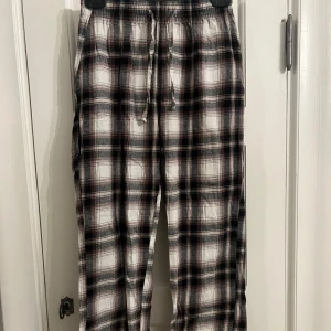 Rutiga pyjamasbyxor - Säljer ett par bekväma rutiga pyjamasbyxor i svart, vit och brun färg. Byxorna har en elastisk midja med snörning för justerbar passform. Perfekta för en avslappnad stil.