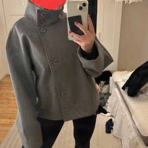 Grå kappa med hög krage - Snygg grå kappa med hög krage och stora knappar. Jackan har en loose passform och långa ärmar, perfekt för vinter/vår. Den stilrena designen gör den lätt att matcha med olika outfits. Älskar den! Är själv 170 och brukar ha M för referens. 