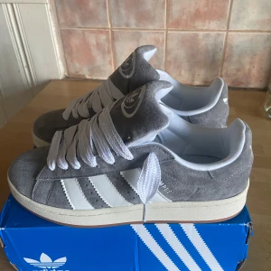 Grå Adidas Campus sneakers - Snygga grå Adidas Campus sneakers i mocka med vita detaljer och snörning. Skorna har den klassiska tre-randiga designen på sidorna och Adidas-loggan på plösen och hälen. Perfekta för en stilren och bekväm look.