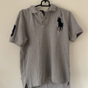 Grå pikétröja från Ralph Lauren - Säljer en klassisk grå pikétröja från Ralph Lauren med korta ärmar. Tröjan har en stor broderad logga på bröstet och en siffra på ärmen. Perfekt för en stilren och avslappnad look. Passar ungefär till 12-14 åringar. Prisa kan diskuteras !