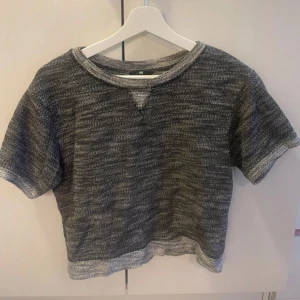 Gråmelerad kortärmad tröja/magtröja  - Säljer en gråmelerad kortärmad tröja med rund hals och avslappnad passform. Perfekt för en casual look. Tröjan har en ribbad kant vid halsen.