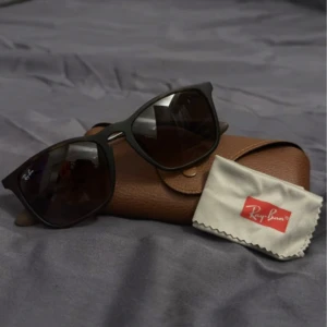 Brunsvarta solglasögon från Ray-Ban - Snygga svarta solglasögon från Ray-Ban med klassisk design. De har en robust båge och mörka linser som ger ett stilrent intryck. Perfekta för soliga dagar och kommer med ett brunt fodral och putsduk.