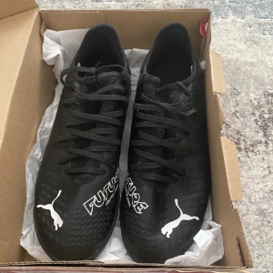 Svarta fotbollsskor från Puma - Snygga svarta fotbollsskor från Puma med texten 'FUTURE' och d Puma-loggan i vitt. Skorna har en strukturerad sula för bra grepp och snörning för optimal passform. Perfekta för fotbollsplanen! Köpt för 500 kr , använt en gång inomhus 