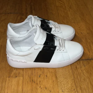 Valentino Garavani sneakers i vitt och svart - Snygga Valentino Garavani sneakers i vitt skinn med en bred svart rem över ovansidan. Skorna har snörning och en stilren design med subtila nitar på hälen. Perfekta för en trendig look. Använda ett fåtal gånger och är i mycket bra skick. Priset är ej hugget i sten så var inte rädda att ställa frågor😁