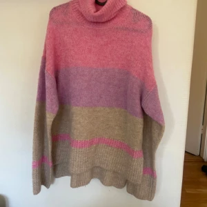 Stickad polotröja - Säljer en mysig stickad polotröja i rosa, lila och beige. Tröjan har en oversized passform med långa ärmar. Aldrig använd så den är i perfekt skick. Köpt på Gekås för nåt år sedan
