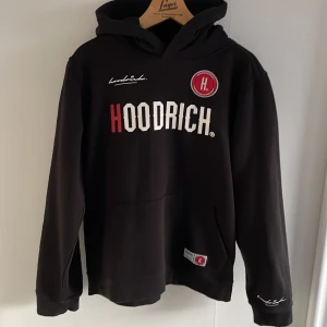 Svart hoodie från Hoodrich - Säljer en svart hoodie från Hoodrich med tryck i vitt och rött på både fram- och baksidan. Hoodien har en stor ficka framtill och en bekväm huva. Perfekt för en avslappnad stil. Tröjan är aldrig använd.                                        Svarar snabbt på frågor!