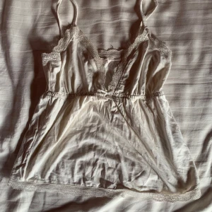 Vit spetsprydd linne från Nelly - Säljer en söt vit linne från Nelly med spetsdetaljer längs halsringningen och en liten rosett framtill. Perfekt för en romantisk look. Linnet har smala axelband och en mjuk, bekväm passform. Kan passa xxs-s!