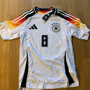 Tyskland fotbollströja - Säljer en vit fotbollströja från Adidas med svarta och röda detaljer. Tröjan har korta ärmar och ett tryck med nummer 8 på framsidan och baksidan. Perfekt för fotbollsfans!