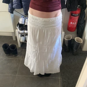 Vit långkjol - Säljer en vit långkjol med elastisk midja. Kjolen har en luftig och bekväm passform, perfekt för varma dagar. Den är tillverkad i ett lätt material som ger en skön känsla. Passar bra till en avslappnad stil.