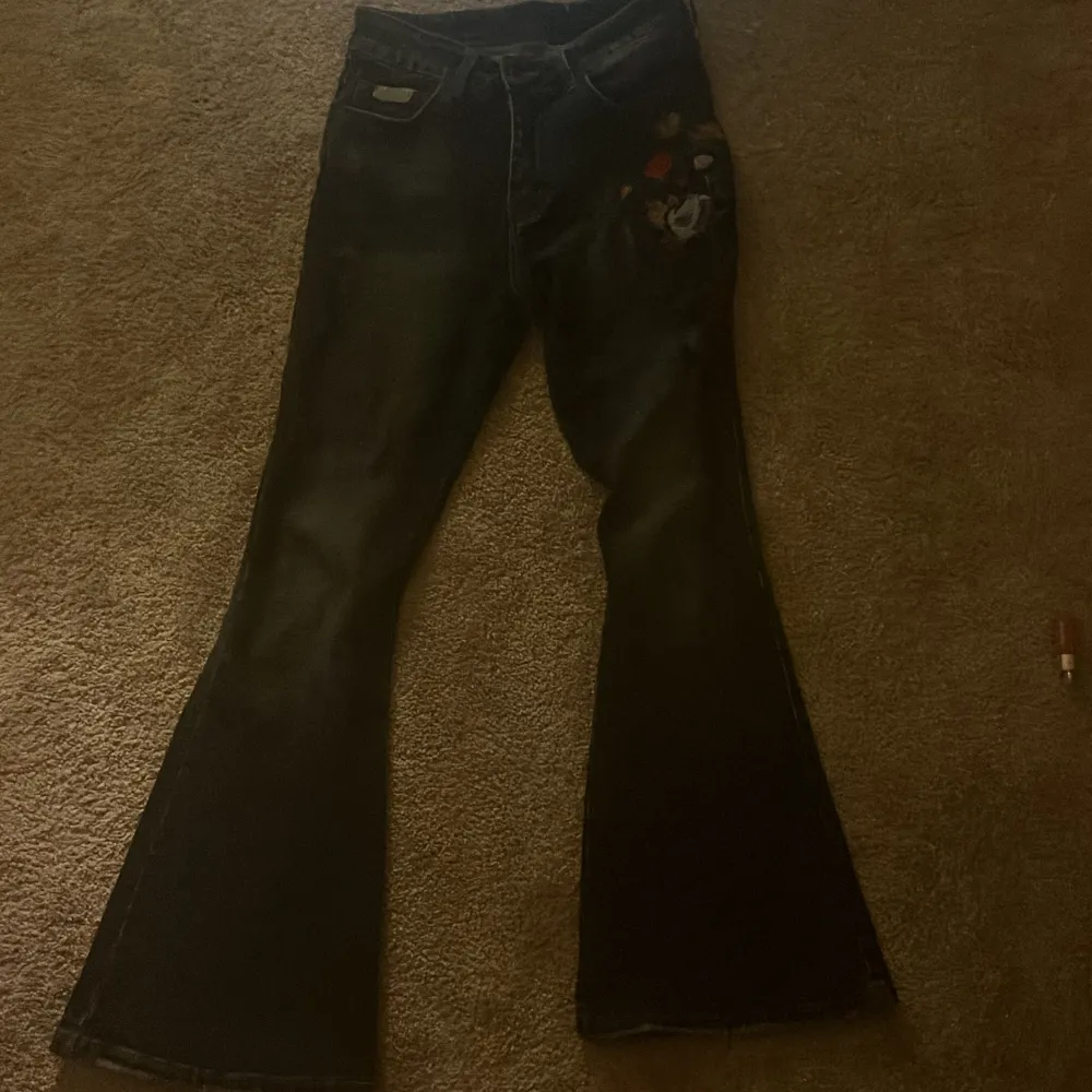 Snygga svarta bootcut jeans med färgglatt blommigt broderi på ena benet. Jeansen har en klassisk femficksdesign och är tillverkade i ett robust denimtyg. Perfekta för att ge din outfit en unik touch.. Farkut & Housut.