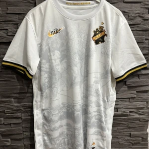  AIK t shirt, Nike - Snygg vit AIK fotbollströja Nike med gulddetaljer och klubbens emblem på bröstet. Tröjan har ett subtilt mönster och korta ärmar med svarta och gula ränder. Perfekt för AIK-fans som vill visa sitt stöd med stil.