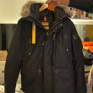 Svart parkajacka från Parajumpers - Säljer en svart parkajacka från Parajumpers med pälsdetaljer på huvan och en gul rem vid kragen. Jackan har flera fickor och en robust dragkedja. Perfekt för kalla dagar! 🖤