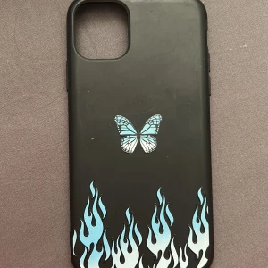 Svart mobilskal med fjäril och flammor (iPhone 11 PRO Max) - Snyggt svart mobilskal med en blå fjäril och coola blå flammor. Perfekt för att ge din telefon en unik stil. Passar till iPhone-modeller.