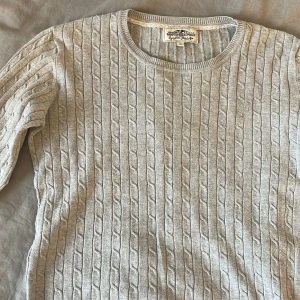 Grå stickad tröja från Hampton Republic - Säljer en stilren grå stickad tröja från Hampton Republic. Tröjan har ett klassiskt kabelstickat mönster och rund halsringning. Perfekt för kyligare dagar med sina långa ärmar och mjuka material. Passar både till jeans och kjol för en avslappnad look.