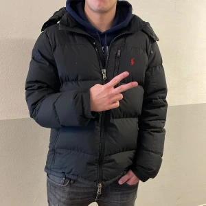 Svart dunjacka med huva - Säljer en svart dunjacka från Ralph Lauren med huva och dragkedja. Jackan har en broderad logga på bröstet och är perfekt för kyliga dagar. Den är långärmad och har en pufferdesign som ger extra värme.
