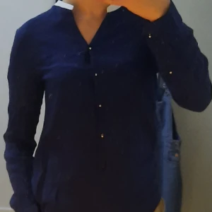 Mörkblå blus från Zara - Säljer en elegant mörkblå blus från Zara i 100% viskos. Blusen har en stilren design med långa ärmar och guldiga knappar framtill samt en vit krage. Perfekt för en chic look.