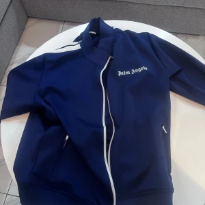 Blå jacka från Palm Angels - Säljer en snygg blå jacka från Palm Angels med vita detaljer. Jackan har dragkedja och långa ärmar med vita ränder längs sidorna. Storlek L funkar M 