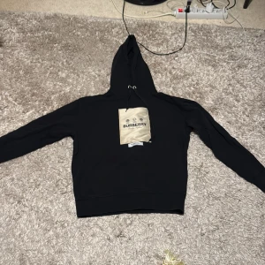 Svart hoodie från Burberry - Snygg svart hoodie från Burberry med en beige patch på framsidan. Hoodien har en klassisk design med dragsko i huvan och ribbade muddar. Perfekt för en stilren look.
