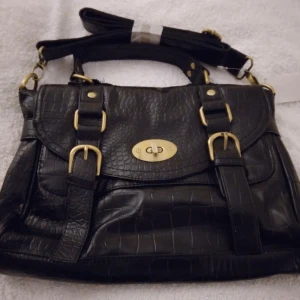 HELT NY & OANVÄND Svart elegant och praktisk väska, krokodil - präglad. - Helt ny och oanvänd svart, praktisk handväska med krokodilskinnsmönster. Klassisk, elegant stil. Slutsåld, från Lindex. Väskan har ett lock med ett vridlås i guldfärgad metall. Det finns två dekorativa remmar med spännen på framsidan. Väskan har ett handtag och en lång, avtagbar och justerbar axelrem. Flera fack med dragkedjor finns. Helt felfri. Mått: ca 32x25x10 cm. (Längd, höjd, djup) Skickas inplastad i originalförpackning.