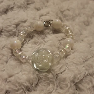 Vitt armband med rosdetalj - Charmigt armband med vita pärlor och en vacker rosdetalj i mitten. Armbandet har en glittrig effekt tack vare de genomskinliga pärlorna som reflekterar ljus. Perfekt för att ge en elegant touch till din outfit.