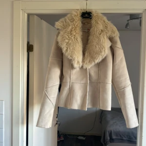 Beige mockajacka med pälsdetaljer - Säljer en superfin beige mockajacka med fluffig päls runt kragen. Jackan har en snygg och stilren design med långa ärmar och en figurnära passform. Perfekt för höst och vinter när du vill hålla dig varm och se chic ut. Passar både till vardags och festliga tillfällen!