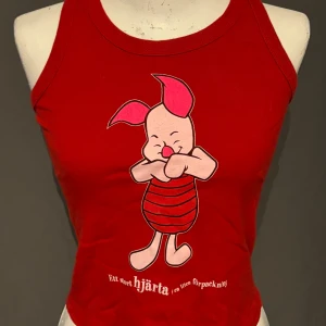Röd ärmlös topp med Nasse-motiv - Säljer en superfin röd ärmlös topp med ett gulligt Nasse-motiv från Nalle Puh. Toppen har texten 'Ett stort hjärta (i en liten förpackning)' tryckt längst ner. Perfekt för sommardagar eller som ett roligt inslag i garderoben!