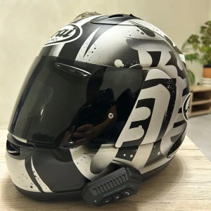 Arai Hjälm med Bluetooth-funktion -  Jag säljer min Arai motorcykelhjälm i storlek Large. Hjälmen är vit med svarta och gråa mönster och har en integrerad Bluetooth-funktion som gör det enkelt att lyssna på musik eller ta emot samtal medan du kör. Den är i mycket gott skick.