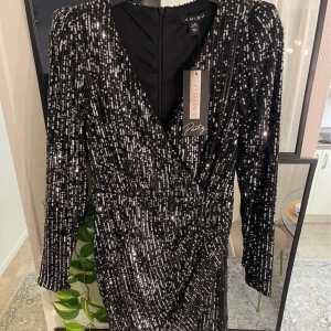 Svart glittrig klänning från Amisu - Säljer en superfin svart glittrig klänning från Amisu. Klänningen har långa ärmar och en snygg omlott-design framtill. Perfekt för festliga tillfällen med sin glittriga look. Den har en dragkedja. 
