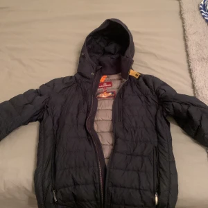 Svart dunjacka från Parajumpers - Säljer en snygg och varm svart dunjacka från Parajumpers. Jackan har en huva och dragkedja framtill. Perfekt för kalla dagar och har en cool design med märkets logga på insidan. Den är i bra skick och redo för nya äventyr! Kom privat för mer frågor 