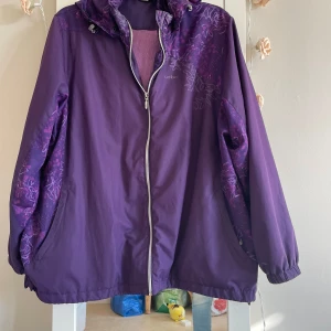 Lila vindjacka från Umbro - Säljer en snygg lila vindjacka från Umbro. Den har ett coolt blommönster i en mörkare nyans av lila och en praktisk dragkedja framtill. Jackan är perfekt för vår och höst med sin lätta och bekväma passform. Den har långa ärmar och en justerbar huva för extra skydd mot vinden.