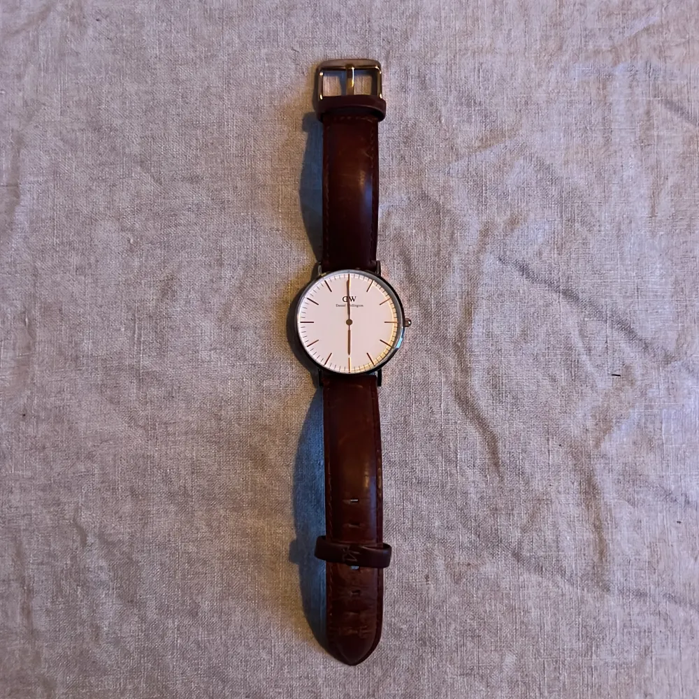 Säljer en stilren klocka från Daniel Wellington med en vit urtavla och brunt läderarmband. Klockan har en minimalistisk design med en tunn boett i champagnefärg. Perfekt för både vardag och fest!. Accessoarer.