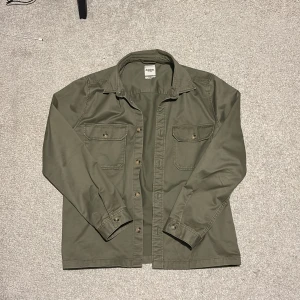 Olivgrön overshirt från Dobber - Säljer en snygg olivgrön overshirt från Dobber i storlek medium. Den har en klassisk krage och knappar framtill. Två bröstfickor med knappar ger en cool look. Perfekt för lager-på-lager-stil under hösten. Skicket är riktigt bra, nästan som ny!