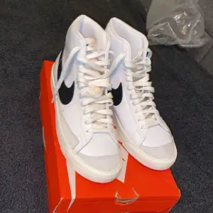 Säljer ett par fräscha vita Nike sneakers med den klassiska svarta swoosh-loggan på sidan. Skorna har en hög modell med snörning och är i nyskick. Kan gå ner i pris vid snabbköp 