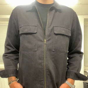 Marinblå overshirt från H&M - Säljer en stilren marinblå overshirt från H&M. Den har en dragkedja framtill och två praktiska bröstfickor med lock. Perfekt för lager-på-lager under hösten eller våren. Materialet är av bomull.